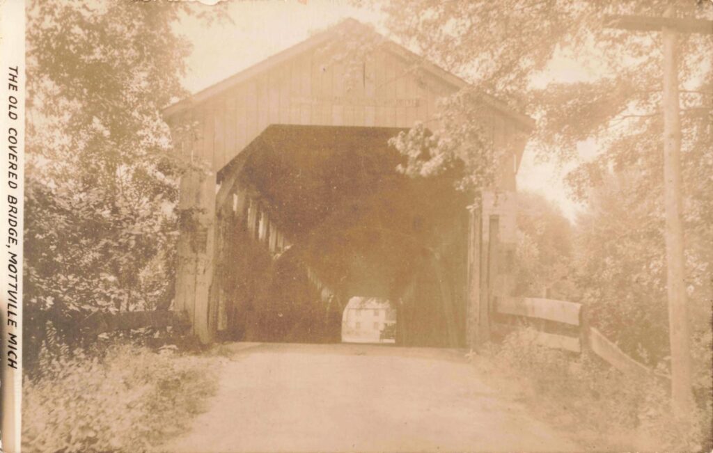 Sepia postcard view of a wooden covered bridge over a dirt road, surrounded by trees, with “THE OLD COVERED BRIDGE, MOTTVILLE MICH” printed along the left edge.