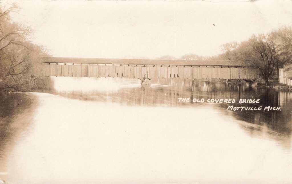 A long wooden covered bridge crosses a river with tree-lined banks, with the words “THE OLD COVERED BRIDGE MOTTVILLE MICH.” written on the image.