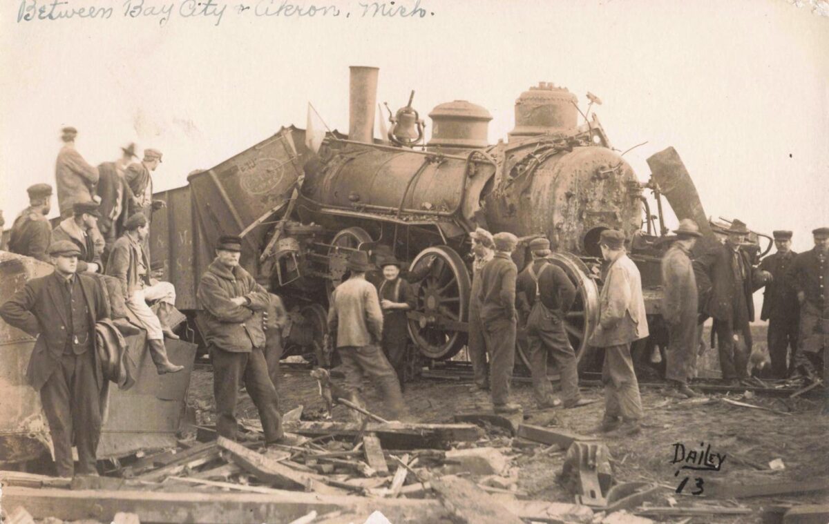 Black-and-white photo of a steam locomotive derailed on its side with a crowd of men standing and watching around the wreckage.