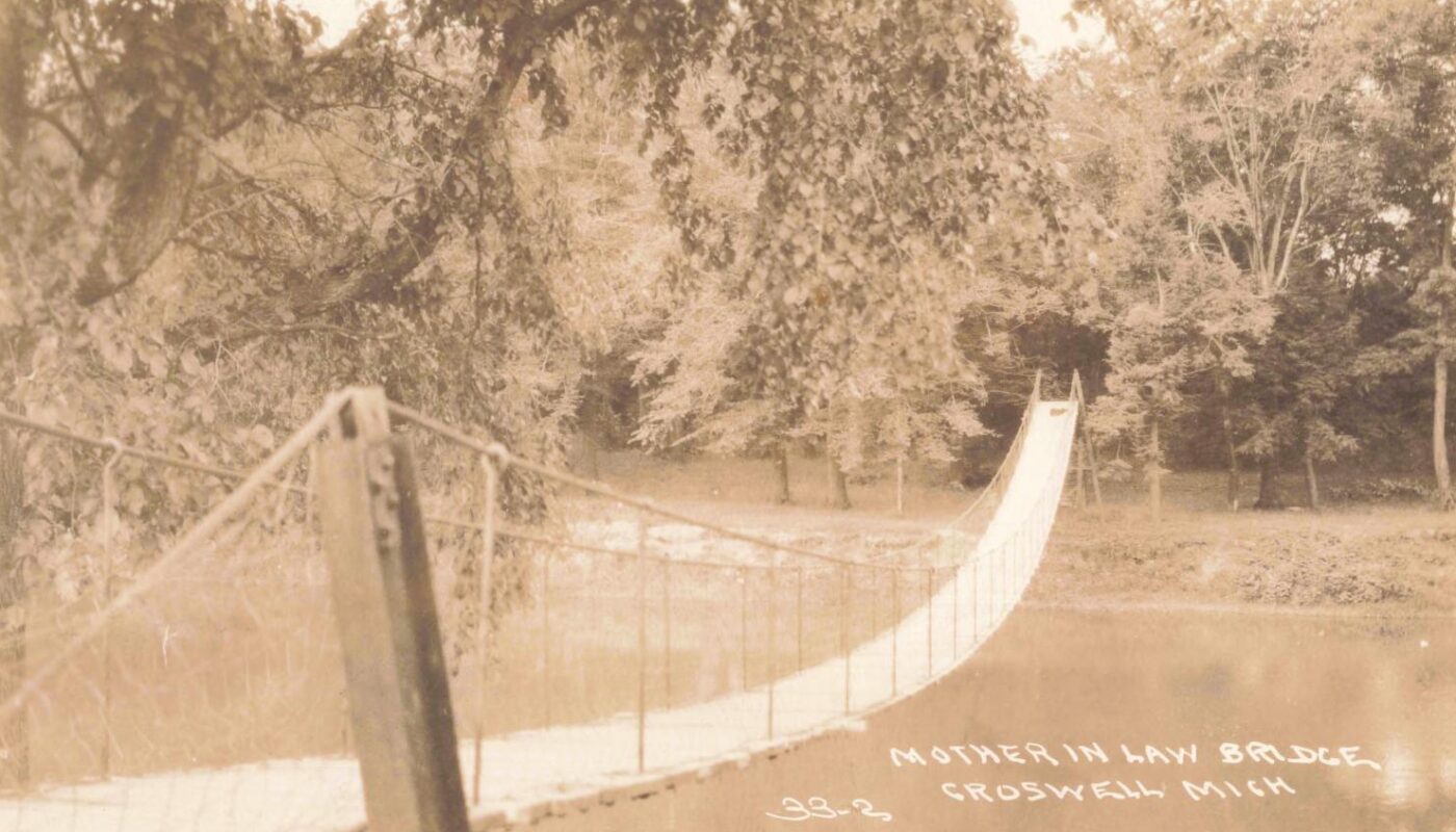 Sepia-toned photo of a narrow suspension footbridge crossing a calm river, with trees and a grassy bank in the background.