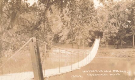 Sepia-toned photo of a narrow suspension footbridge crossing a calm river, with trees and a grassy bank in the background.