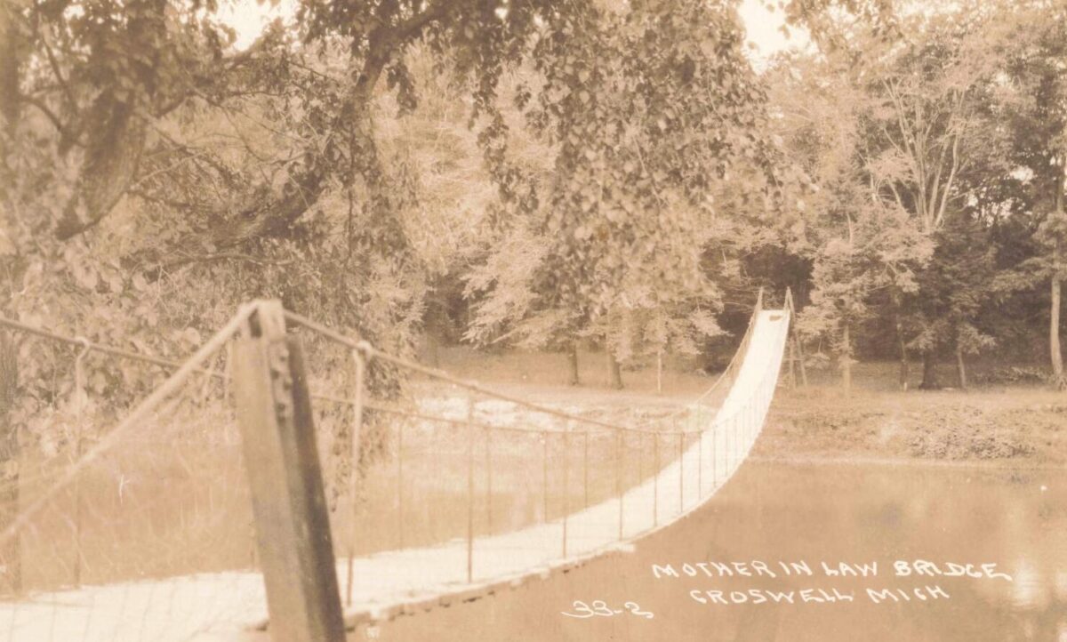 Sepia-toned photo of a narrow suspension footbridge crossing a calm river, with trees and a grassy bank in the background.