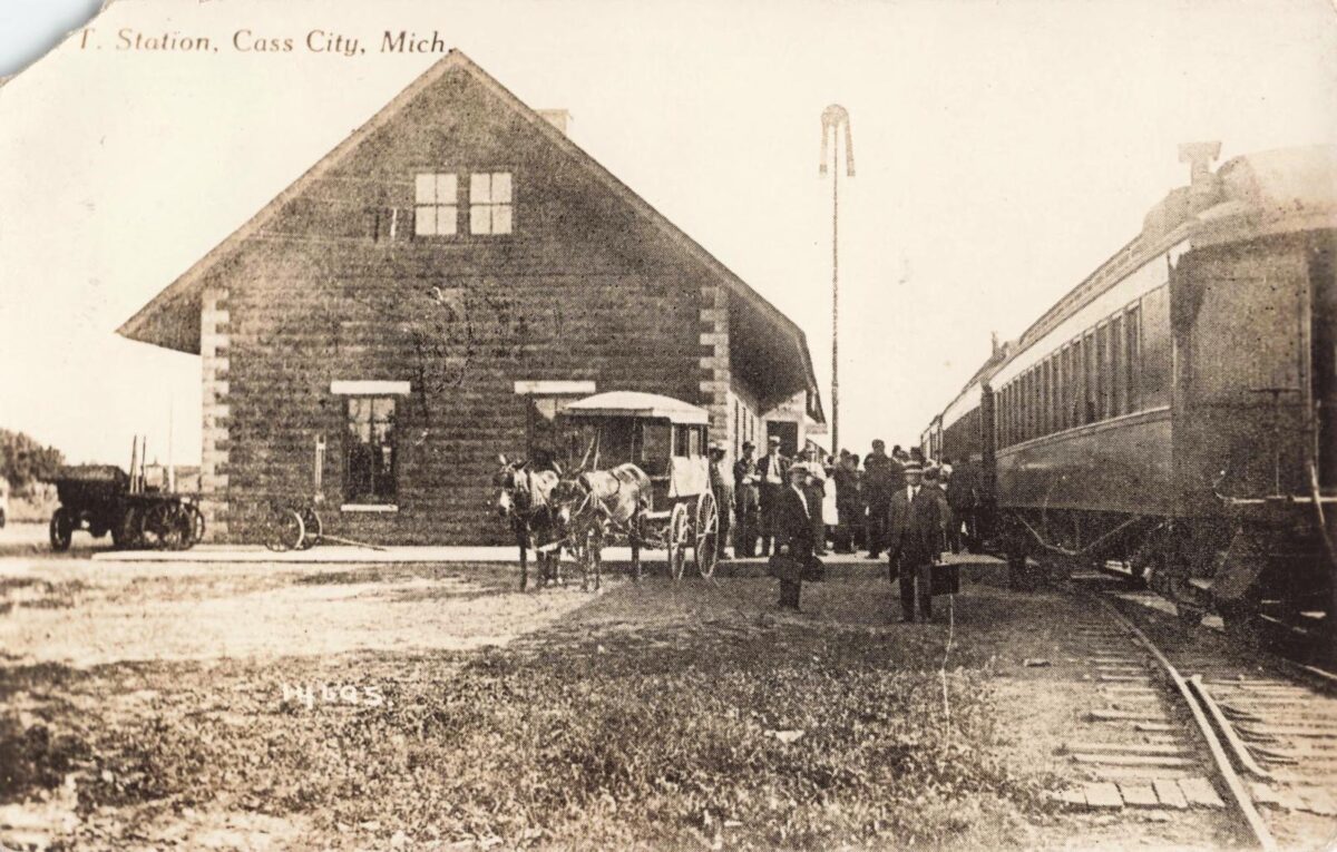 Black-and-white postcard of people boarding a train beside a brick Grand Trunk station in Cass City, Michigan, with a horse-drawn carriage near the depot.