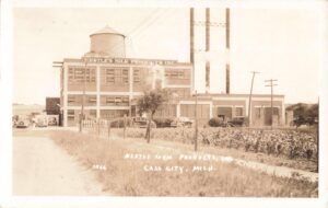 Sepia-toned photo of a large factory labeled “Nestlé’s Milk Products Inc.” with a water tower, three smokestacks, several old cars, and fields in the foreground.
