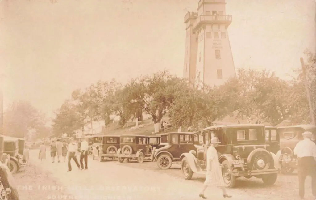 a group of people walking on a road with cars parked in front of a lighthouse