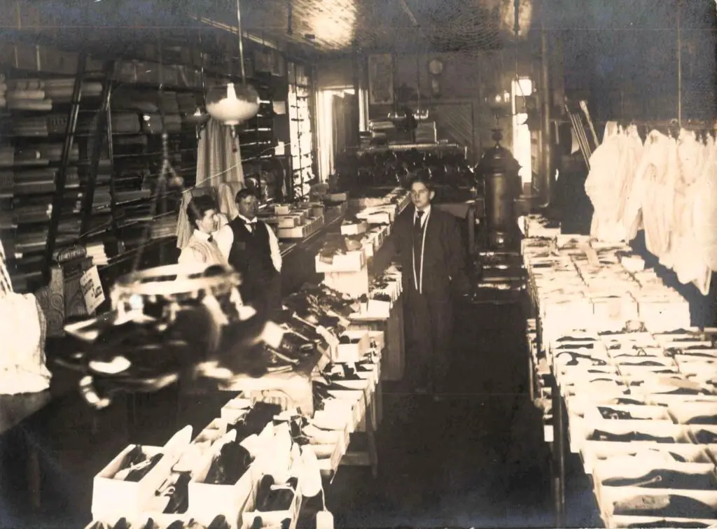Ed Rogers’ Shoe Store: Interior crowded with shoe boxes; clerks under hanging lamps.