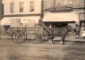 A horse-drawn wagon stacked with wooden crates sits in front of storefronts, including a sign reading “H.C. Clabuesch City Drugstore,” with two children standing near the entrance.