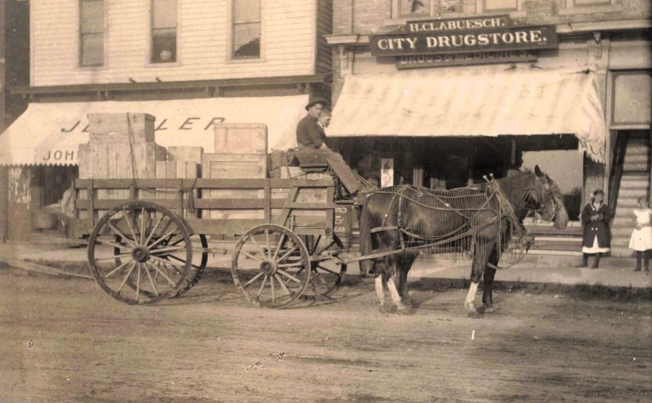 A horse-drawn wagon stacked with wooden crates sits in front of storefronts, including a sign reading “H.C. Clabuesch City Drugstore,” with two children standing near the entrance.