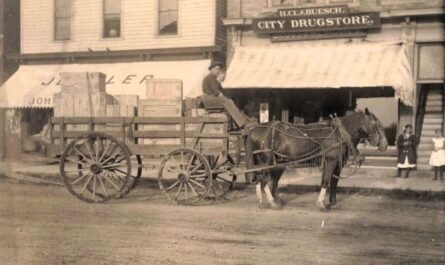 A horse-drawn wagon stacked with wooden crates sits in front of storefronts, including a sign reading “H.C. Clabuesch City Drugstore,” with two children standing near the entrance.