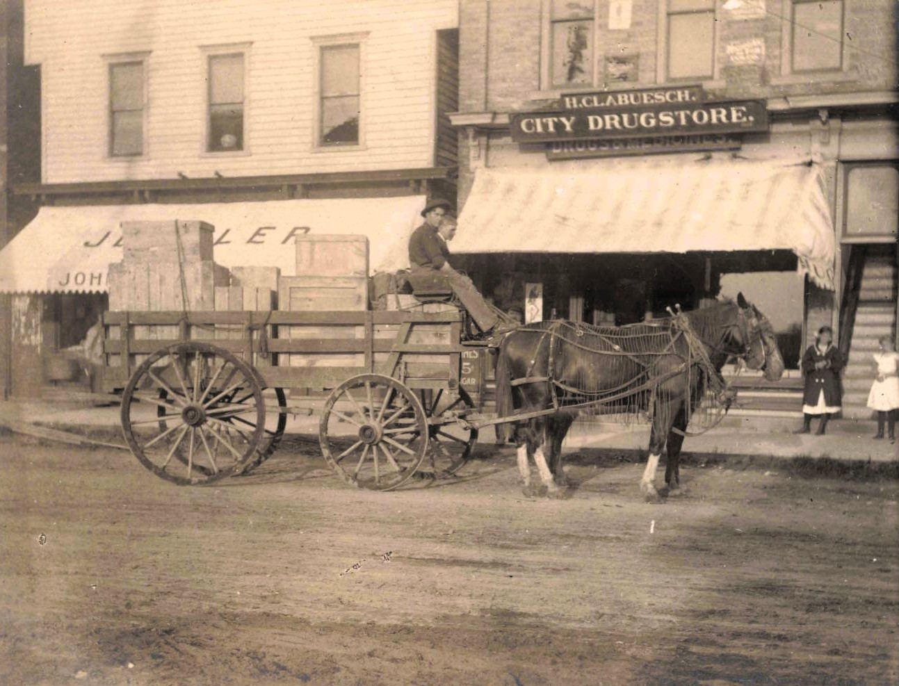 A horse-drawn wagon stacked with wooden crates sits in front of storefronts, including a sign reading “H.C. Clabuesch City Drugstore,” with two children standing near the entrance.