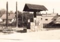 Black-and-white photo of a stone monument with a small roof-like cap, set beside a road, with buildings and business signs in the background; “Sebewaing Mich.” is written at bottom left.