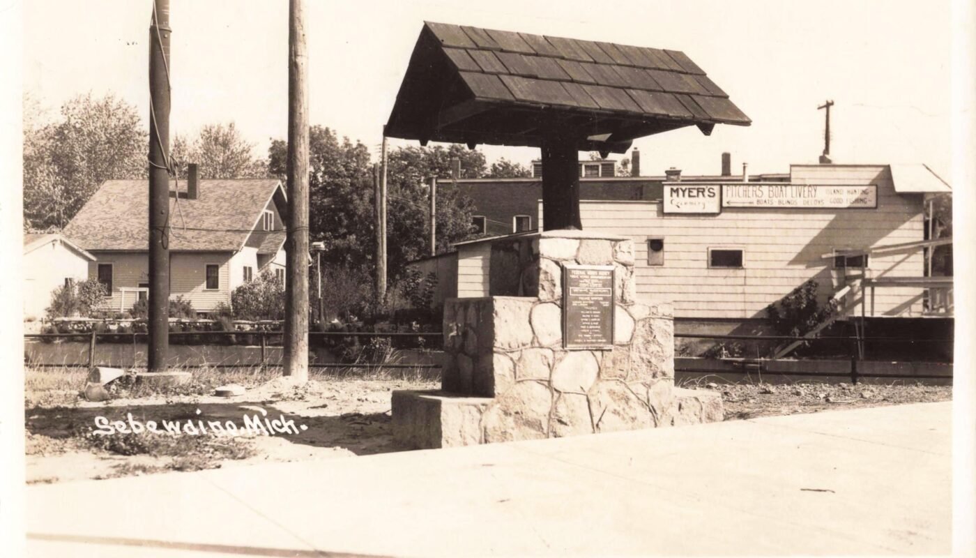 Black-and-white photo of a stone monument with a small roof-like cap, set beside a road, with buildings and business signs in the background; “Sebewaing Mich.” is written at bottom left.