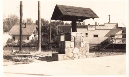Black-and-white photo of a stone monument with a small roof-like cap, set beside a road, with buildings and business signs in the background; “Sebewaing Mich.” is written at bottom left.