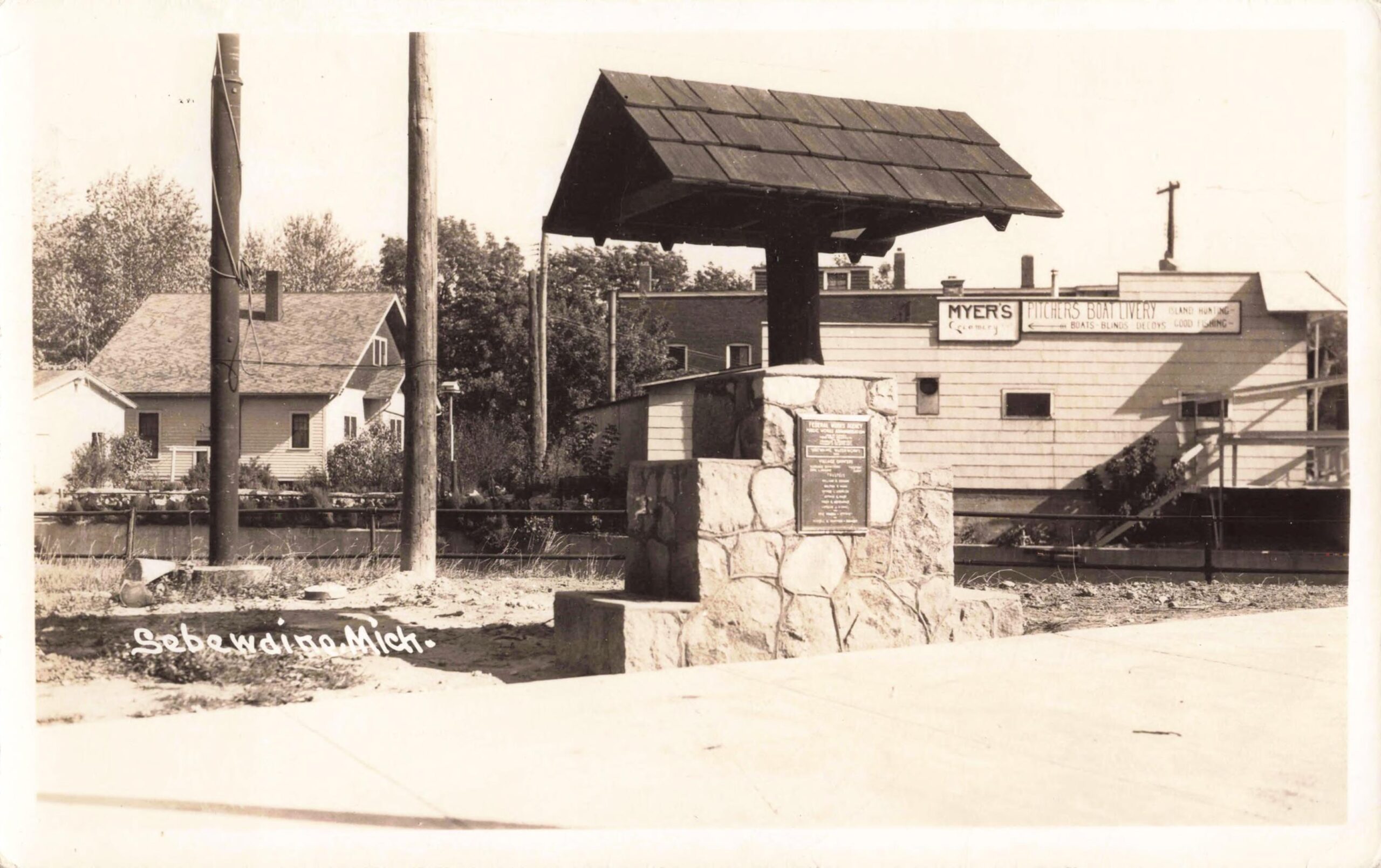 Black-and-white photo of a stone monument with a small roof-like cap, set beside a road, with buildings and business signs in the background; “Sebewaing Mich.” is written at bottom left.