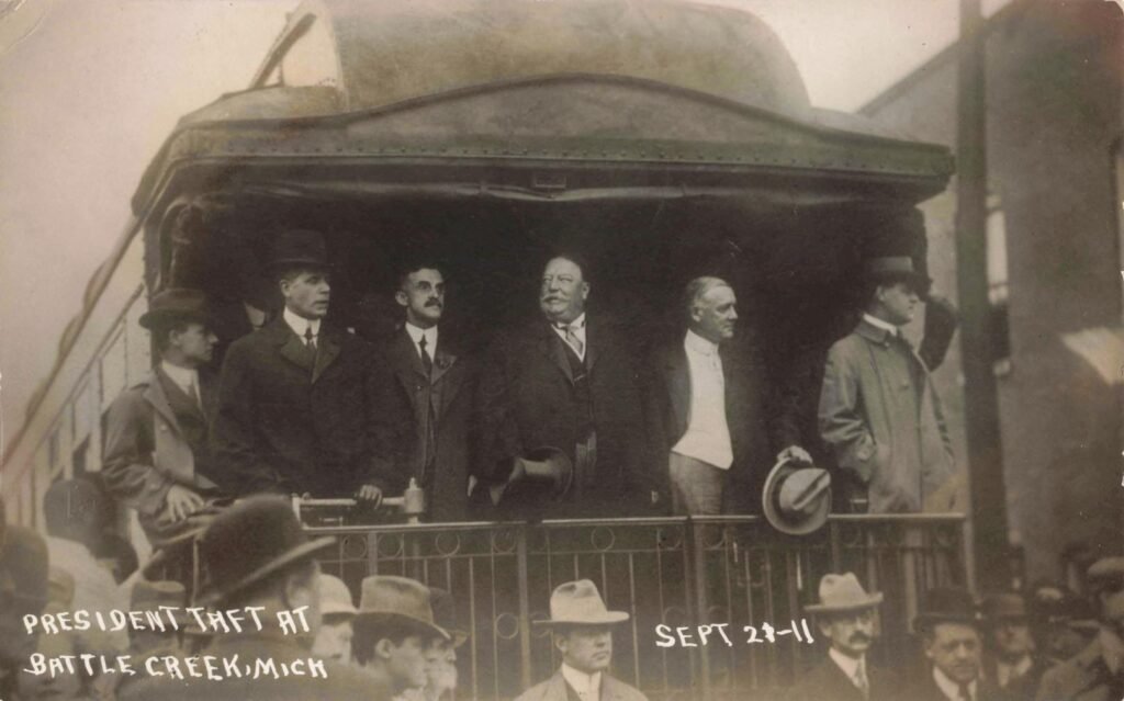 A large crowd watches a man speaking on a platform in front of a building decorated with American flags and bunting, labeled “President Taft at Battle Creek, Mich. Sept. 21, 1911.”