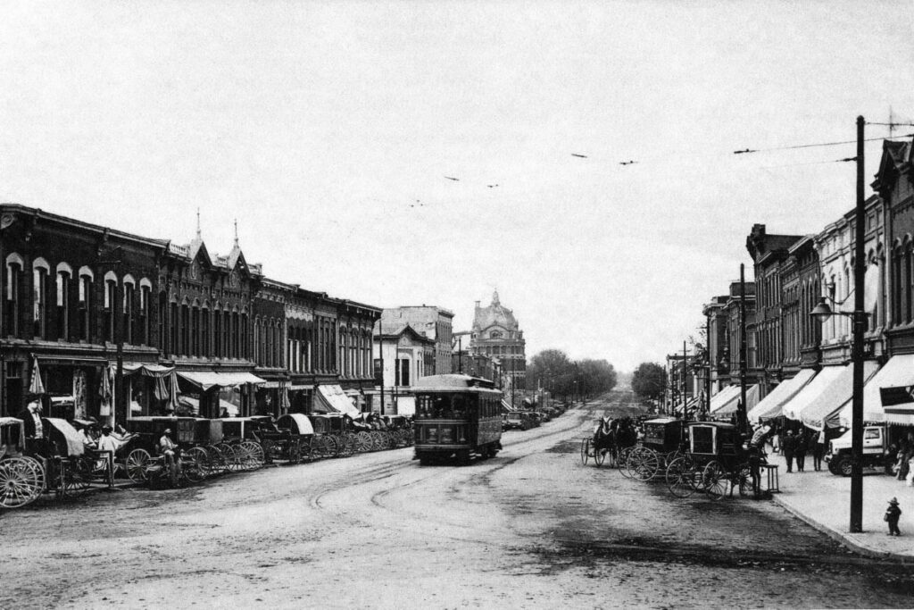 Black-and-white photo of an interurban streetcar in the middle of a wide downtown street with brick buildings on both sides, wagons and bicycles near the curbs, and overhead electric wires.