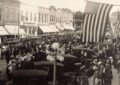 Black-and-white photo of a crowded downtown street with many early cars parked and large American flags hanging overhead, with storefronts lining the block.