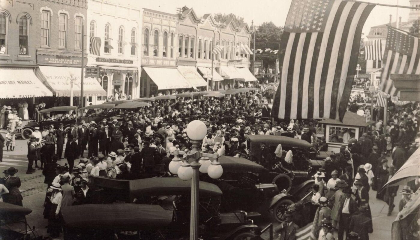 Black-and-white photo of a crowded downtown street with many early cars parked and large American flags hanging overhead, with storefronts lining the block.