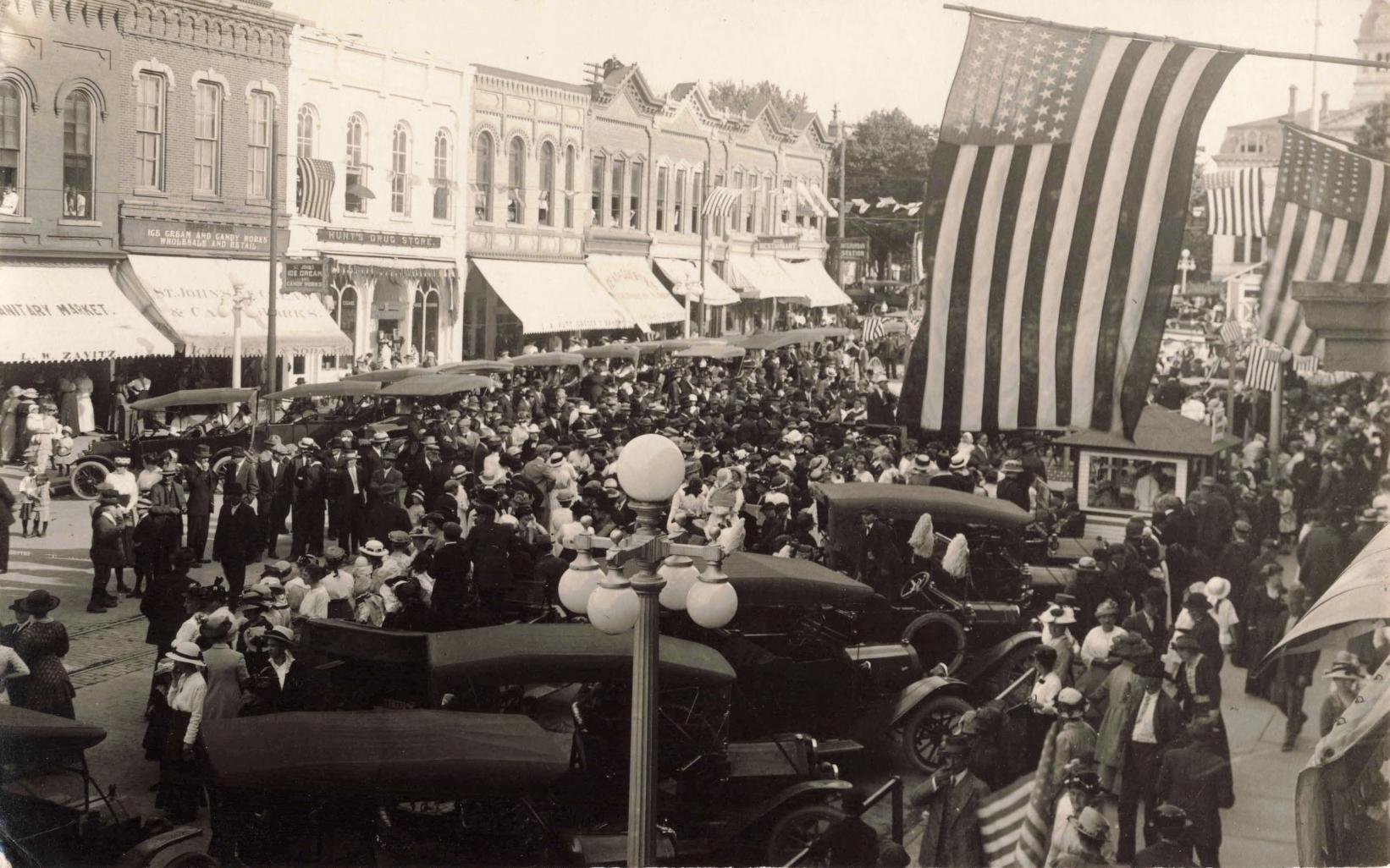 Black-and-white photo of a crowded downtown street with many early cars parked and large American flags hanging overhead, with storefronts lining the block.