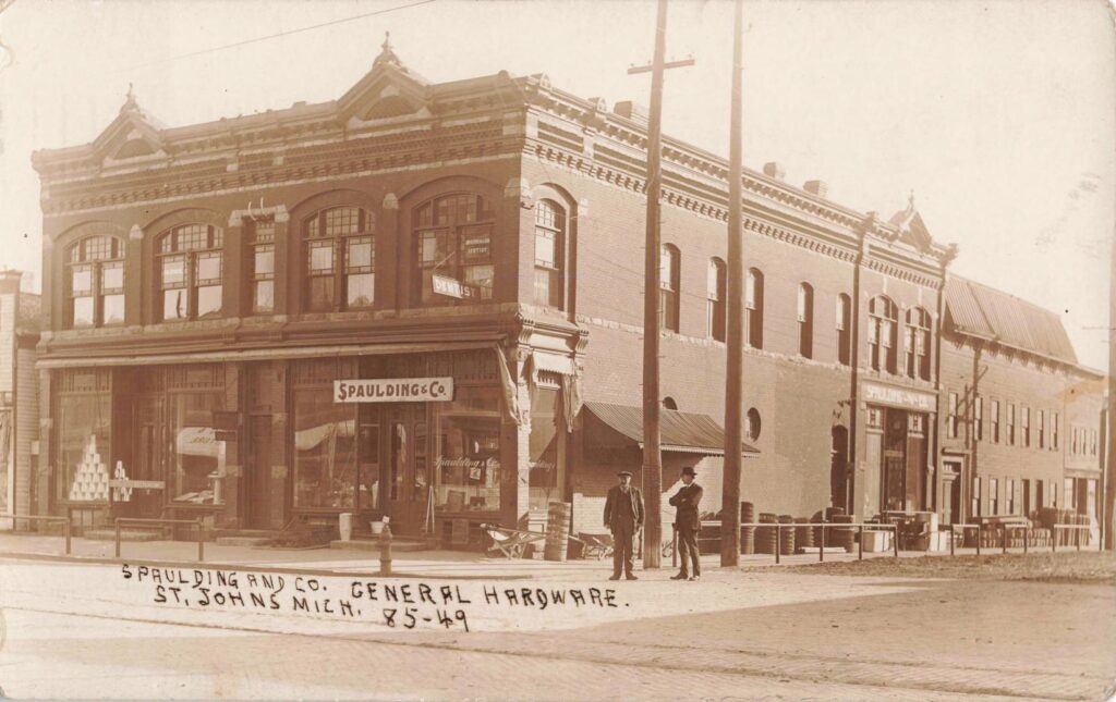 Sepia-toned street view of a two-story brick corner building labeled “Spaulding & Co.” with two men standing near utility poles and a “Dentist” sign in an upstairs window.