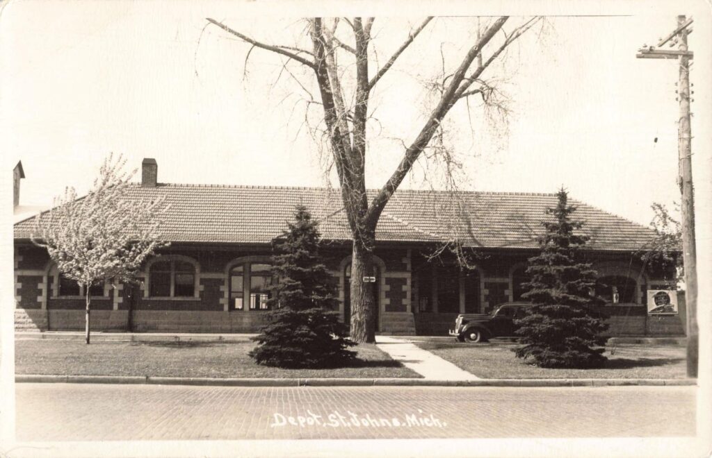 Black-and-white photo of a long, one-story depot building with arched windows and a tile roof, trees in front, a parked vintage car near the entrance, and the caption “Depot, St. Johns, Mich.”