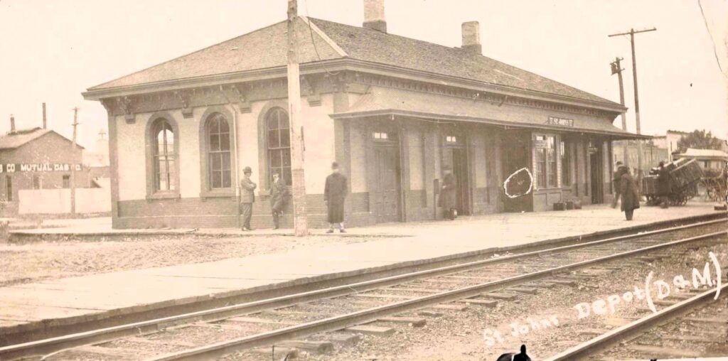 Sepia postcard photo of the St. Johns railroad depot with tracks in front, several people on the platform, wagons at right, and a distant building with “Mutual Gas” signage at left.