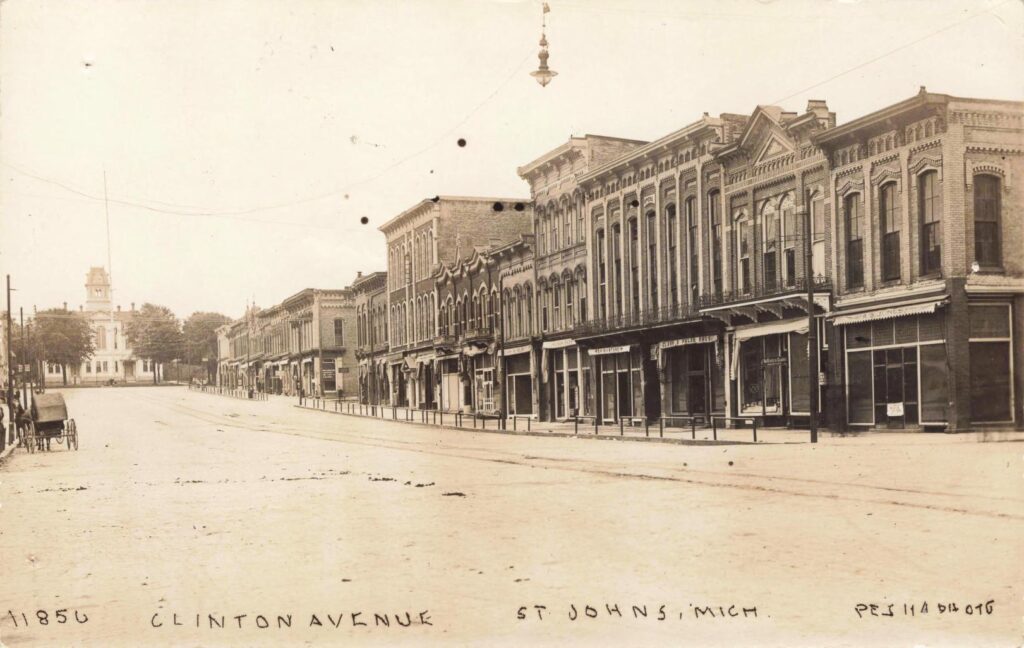 Sepia photo of a wide street with brick storefronts on the right, overhead wires and a hanging streetlight, and a horse-drawn buggy on the left, labeled “Clinton Avenue, St. Johns, Mich.”