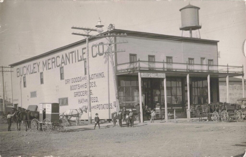 Two-story Buckley Mercantile Co. store with painted signs, a rooftop water tank, and horse-drawn wagons parked on a dirt street.