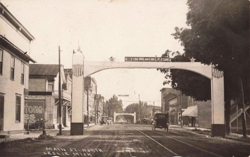 Early 1900s Main Street Leslie Michigan with welcome arch, storefronts, and dirt road traffic.
