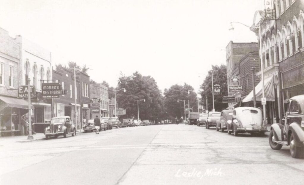 Black-and-white view down a wide Main Street in Leslie, Michigan, with parked vintage cars on both sides and storefront signs including “Morea’s Restaurant” and appliance advertising.