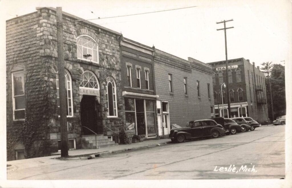 A stone-front building labeled “G.A.R. Hall” stands along a street in Leslie, Michigan, with several vintage cars parked in front and more buildings stretching down the block.