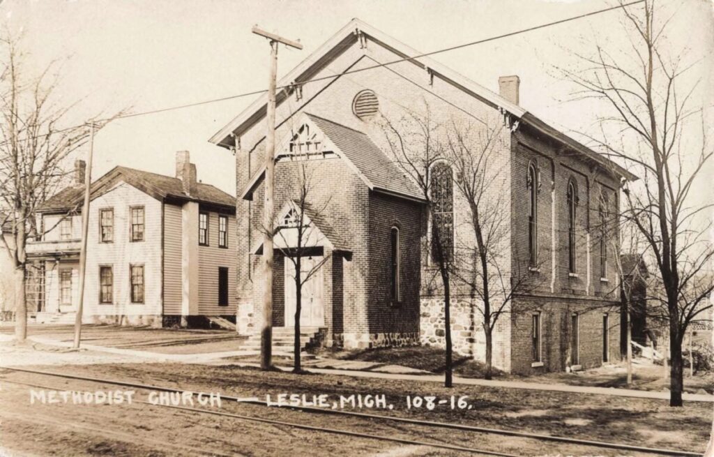 A sepia photo shows a brick church with tall arched windows beside a street, with rail tracks in the foreground and leafless trees around the building.