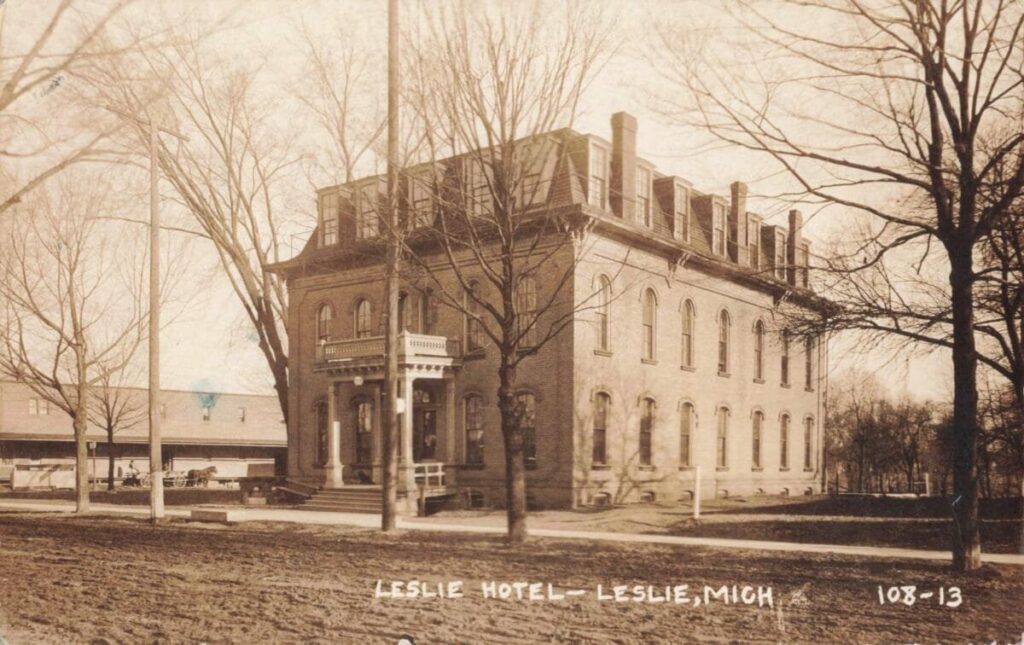 Sepia-toned postcard photo of a large brick hotel labeled “Leslie Hotel — Leslie, Mich,” with a mansard roof, dormer windows, and leafless trees along a dirt street.