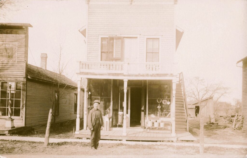 Sepia photo of a man standing on a wooden boardwalk in front of a two-story clapboard storefront with neighboring shops on both sides.