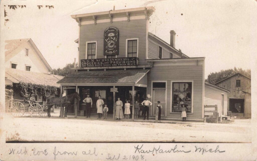 Historic 1908 photo of G.M. Schweinsberg’s general store and post office in Kawkawlin, Michigan. Men, women, and children stand along the wooden porch while a horse-drawn wagon waits at left. A large sign advertises groceries, dry goods, boots, shoes, and fresh meats, with a fraternal lodge sign for Court Kawkawlin No. 3559 (Independent Order of Foresters) mounted above the storefront.
