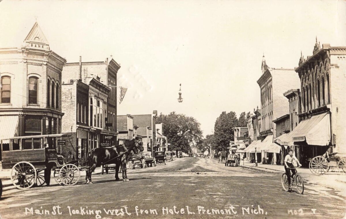 Sepia-toned street scene on Main Street in Fremont, Michigan, showing brick storefronts, a horse-drawn wagon, early cars, and a bicyclist in the foreground.