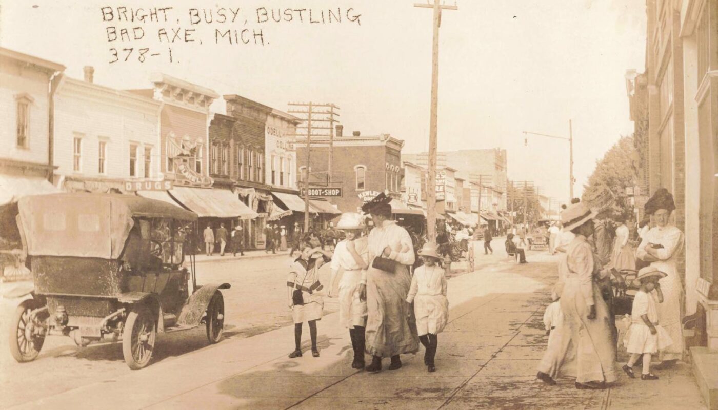 Early 1910s downtown Bad Axe Michigan showing dirt streets, wooden storefronts, wagons, horses, and early automobiles.