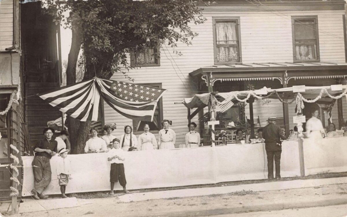 Black-and-white postcard photo of adults and children standing beside a decorated parade display with U.S. flags and bunting outside a wood-frame building.