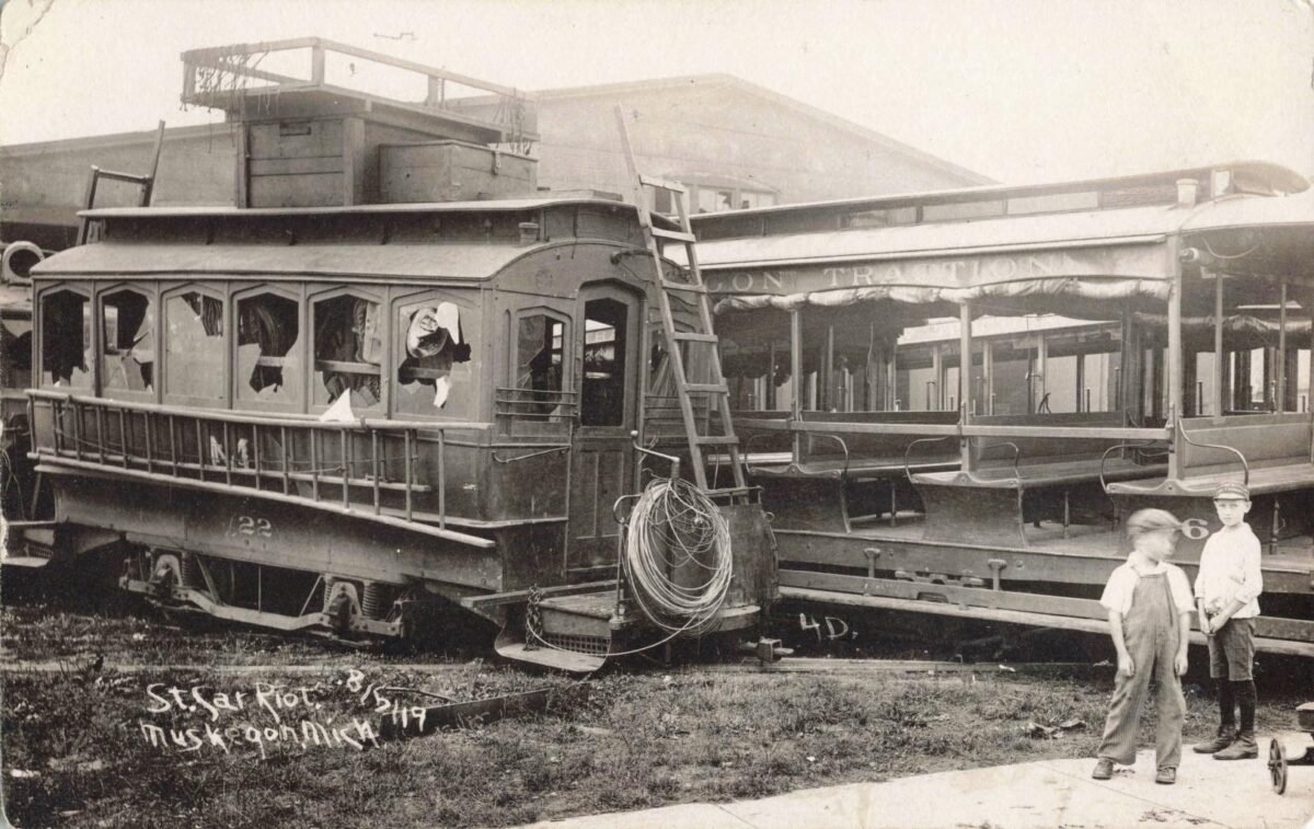 Two damaged 1919 Muskegon streetcars with broken windows and ladders leaning against them, with two boys standing beside the tracks.