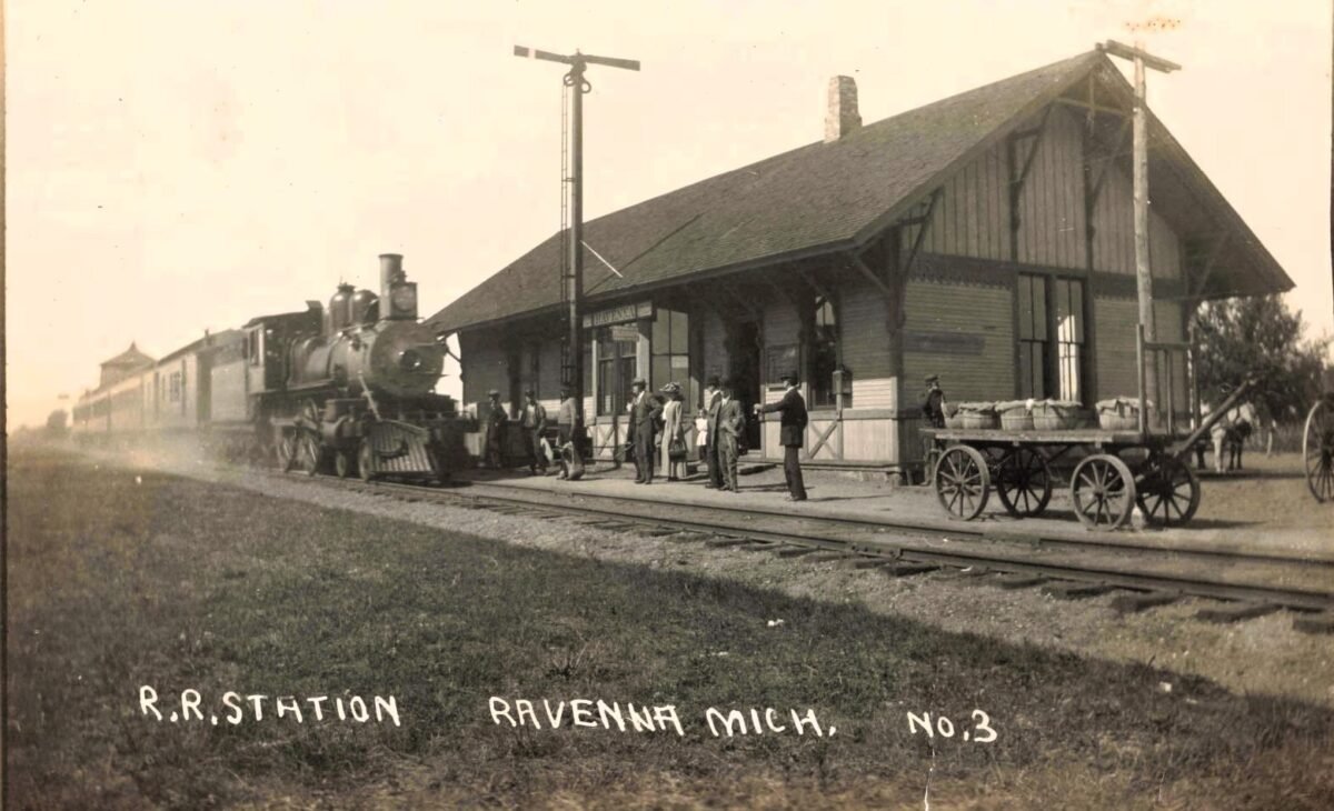 A steam locomotive and passenger cars stop beside Ravenna’s wooden railroad depot as passengers and railroad workers stand on the platform near a horse-drawn wagon.