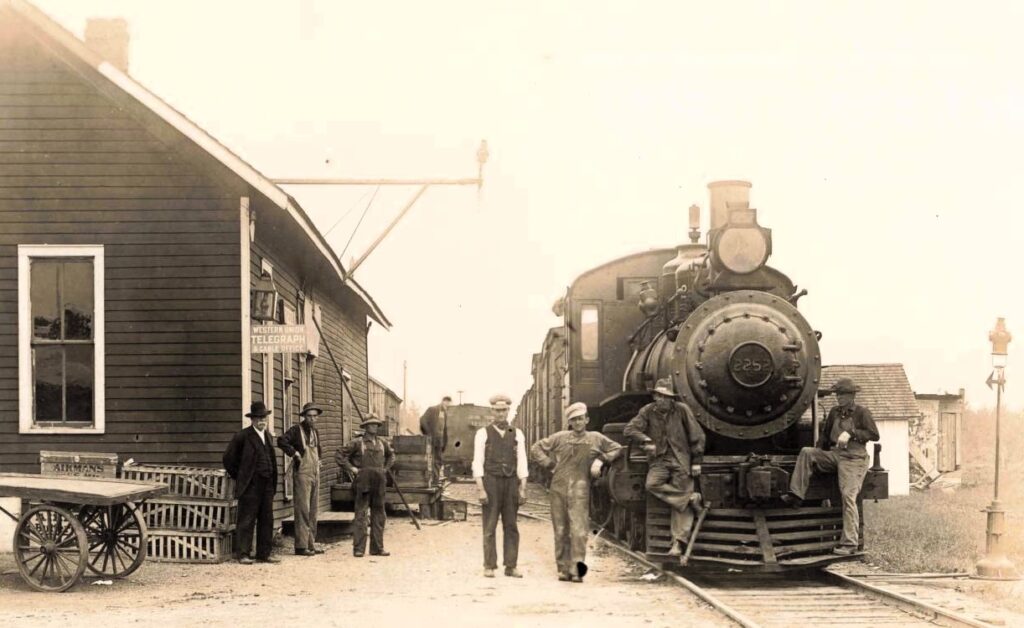 Passengers gather beside the Deford depot as a train prepares for departure. Rail travel connected small towns with Detroit, Pontiac, and other Michigan communities. - History of Deford Michigan
