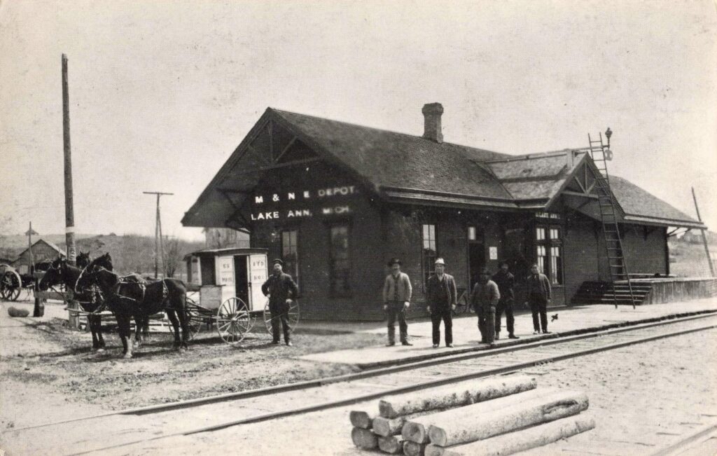 The Manistee & Northeastern depot at Lake Ann, with men, horses, rail tracks and logs showing the village’s link to lumber and transportation.