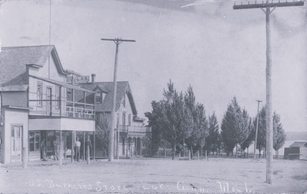 S.S. Burnett’s store in Lake Ann, with early village storefronts, utility poles and the lake visible nearby.