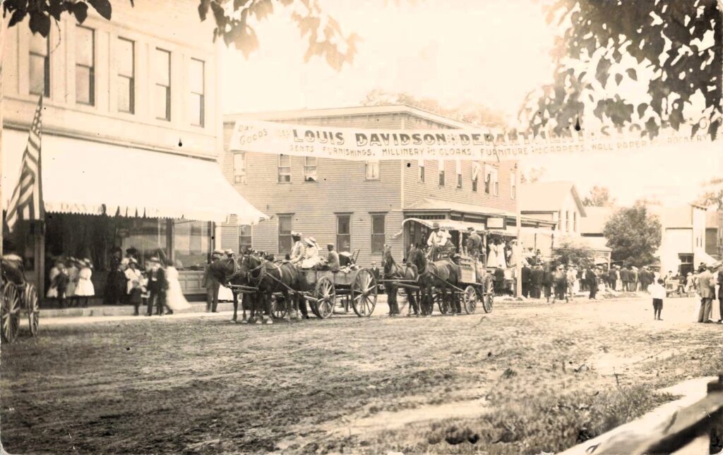 State Street in history of Hillman Michigan, with the Louis Davidson Department Store visible, shows a dirt main street filled with horses, wagons, overhead wires and commercial activity.