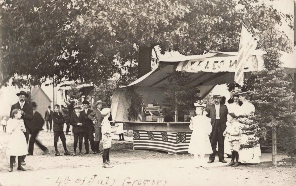 Children and adults standing near a flag-covered Fourth of July booth at a church picnic in Gregory Michigan in 1908