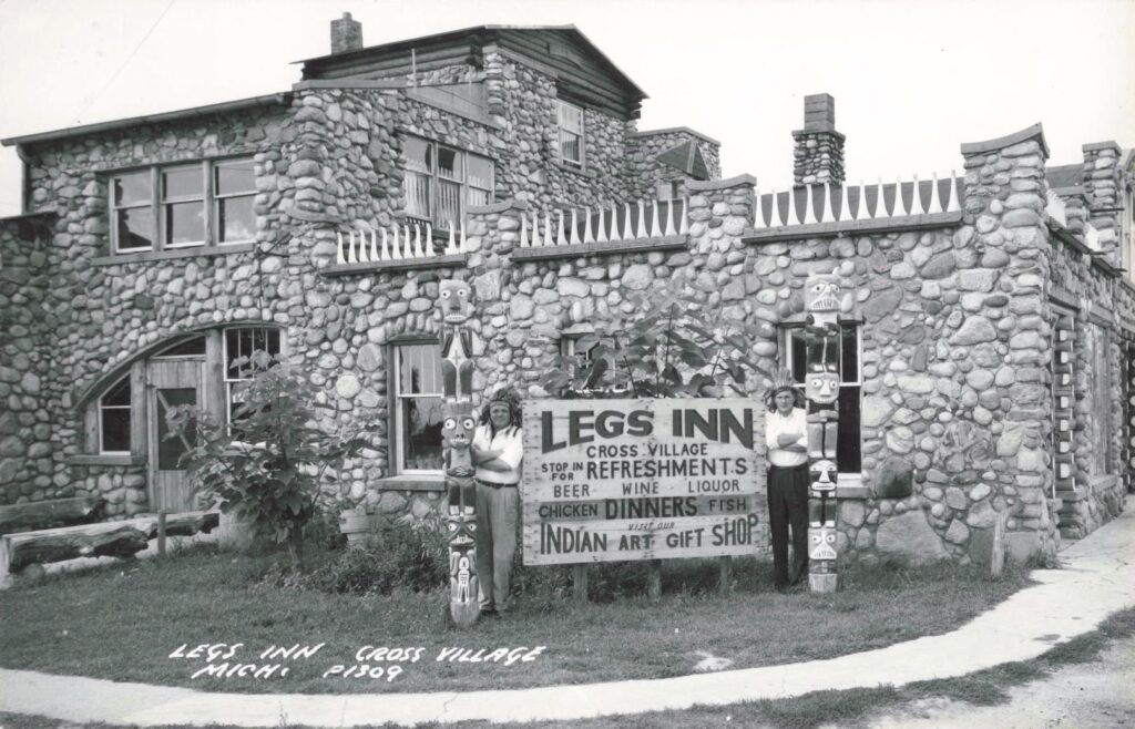 Historic exterior postcard of Legs Inn in Cross Village, Michigan, showing the stone building, stove-leg roofline and carved poles.