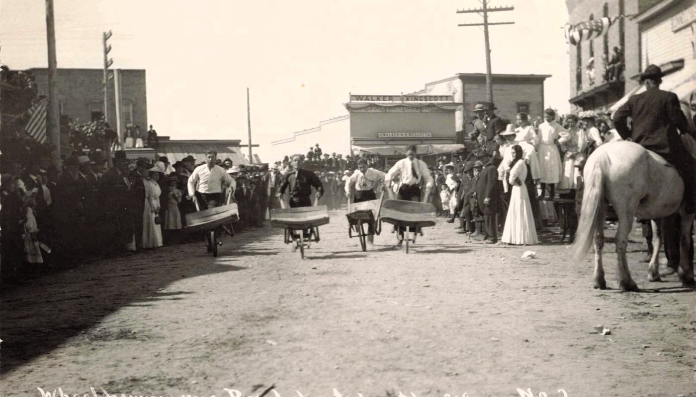 Men push wheelbarrows down a dirt street while crowds watch during a July 4, 1909, celebration in Bear Lake, Michigan.