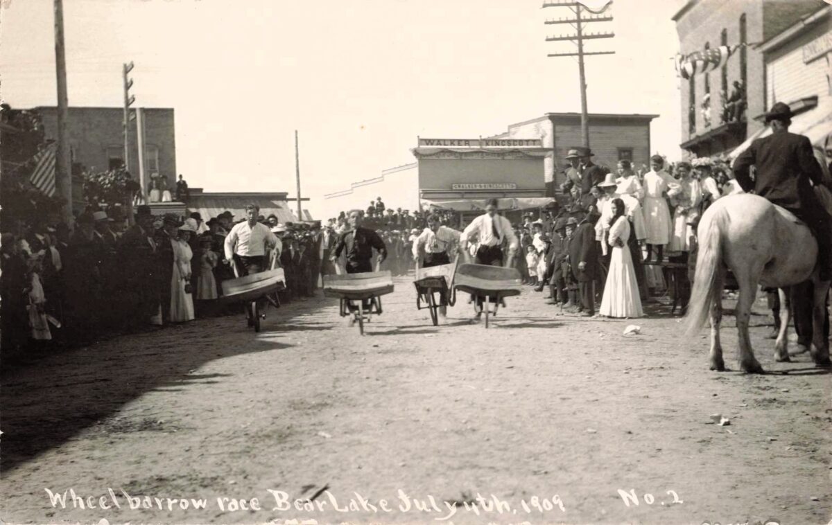 Men push wheelbarrows down a dirt street while crowds watch during a July 4, 1909, celebration in Bear Lake, Michigan.