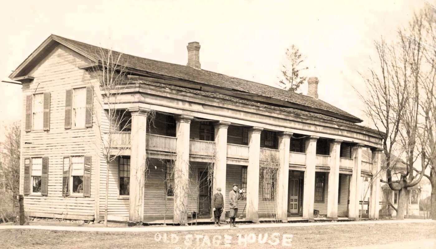Historic two-story wooden Old Stage House in Clinton, Michigan, with a long columned porch and two people standing near the front entrance.