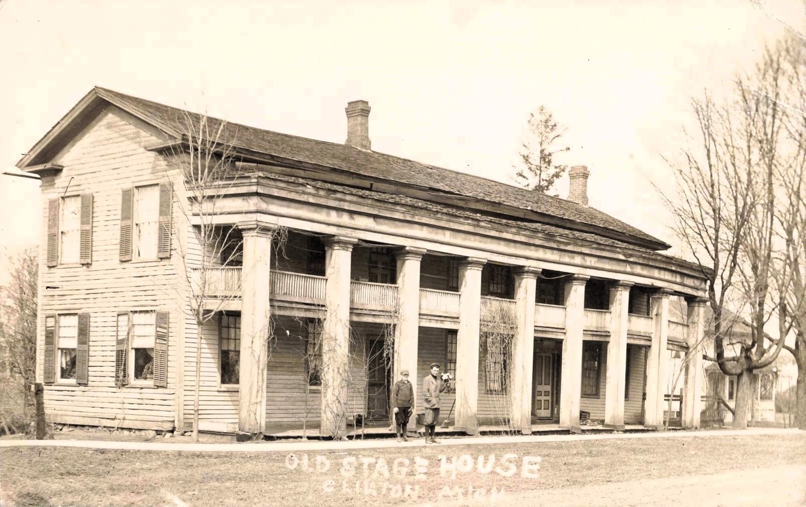 Historic two-story wooden Old Stage House in Clinton, Michigan, with a long columned porch and two people standing near the front entrance.